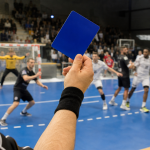 Arbitre de handball levant un carton bleu pendant un match en salle, avec joueurs et but flous en arrière-plan