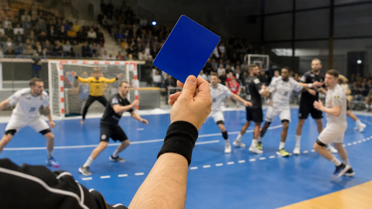 Arbitre de handball levant un carton bleu pendant un match en salle, avec joueurs et but flous en arrière-plan